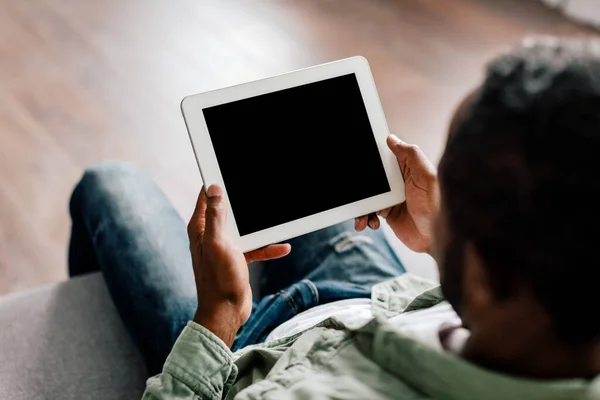 Mature african american male watching video on tablet with empty screen, sit on sofa in living room interior, over shoulder view. New website, app, ad and offer for work at home during free time