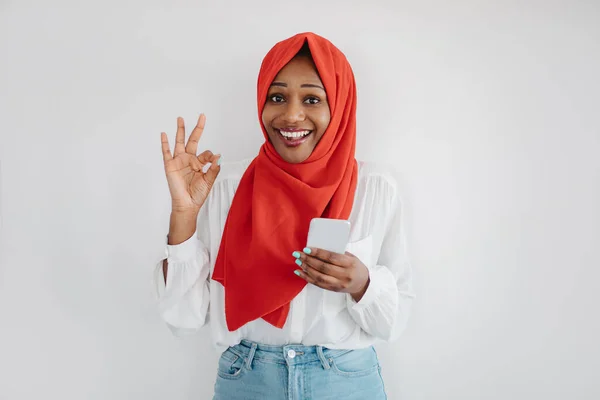 伟大的应用程序 在头巾里兴奋的黑人穆斯林女人使用智能手机 摆出一副好的姿势 在轻墙的背景上摆出姿势 腾出空间 — 图库照片