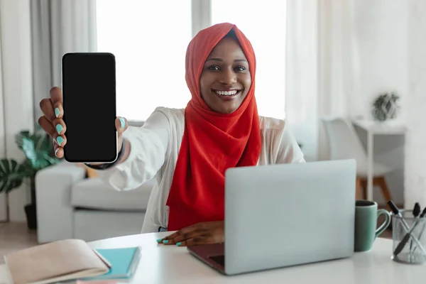 Happy black muslim woman with laptop showing smartphone with blank screen, recommending new mobile app or website at home office, mockup. Modern technologies in everyday life