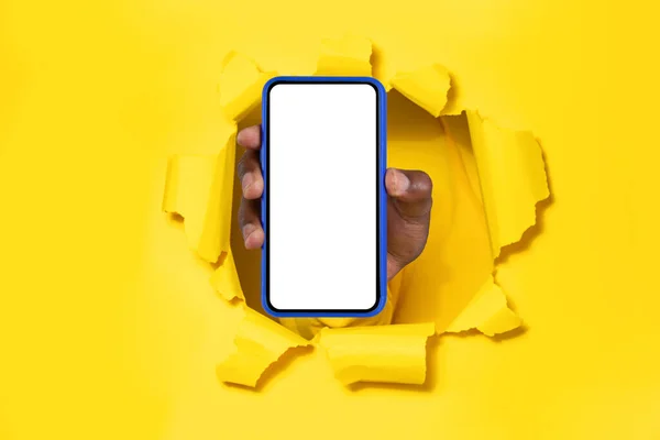 Mobile app promo. Black male hand holding cellphone with white empty screen showing device to camera breaking through yellow paper sheet. Mock up