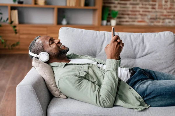 無線ヘッドフォンでカジュアルで陽気な成熟したアフリカ系アメリカ人男性は リビングルームのインテリアにソファの上にあり スマートフォンでお気に入りの音楽を楽しんでいます 自宅で休息とリラックス オーディオアプリ 歌で休憩 — ストック写真