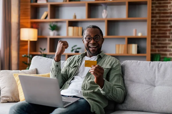 Glad excited adult black man in glasses and casual rejoices in victory with computer and credit card on sofa in living room interior. Online shopping, business and work remotely at home, sale and win