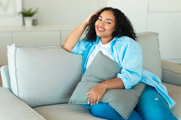 Happy African American Lady Resting Sitting On Sofa Holding Pillow Posing Smiling To Camera At Home. Relaxed Woman Enjoying Domestic Comfort On Weekend. Rest And Relaxation