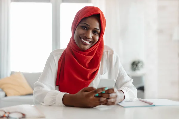 Portrait of happy black muslim businesswoman in hijab using cellphone while sitting at desk and smiling at camera, african american lady texting on smartphone while working in office