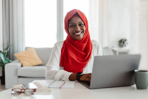 Happy muslim african american woman sitting at workplace and smiling at camera while working on laptop, posing alone at cozy home office, copy space. Millennials business concept