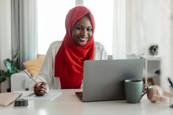 Note taking. Positive muslim african american woman sitting at workplace, using laptop and writing in notebook, home office. Excited black lady studying online from home, remote work