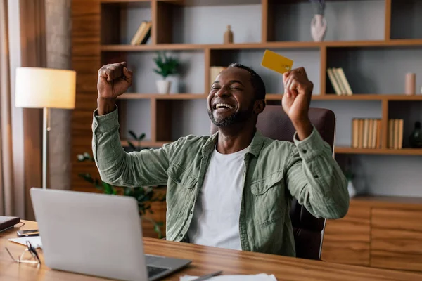 Satisfied excited adult african american man in casual rejoices to winning, cashback and online shopping with laptop and credit card in home office interior. Huge sale, success victory and technology