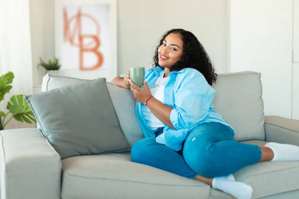Relaxed Black Woman Holding Coffee Cup Smiling To Camera Resting Sitting On Sofa Posing In Modern Living Room At Home. Domestic Comfort And Weekend Leisure Concept