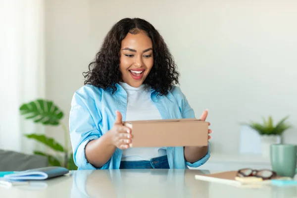 Excited Black Lady Buyer Holding Cardboard Box Unpacking Delivered Parcel With Plus Size Clothes Sitting At Workplace Indoors. Modern Shopping And Delivery Concept. Selective Focus