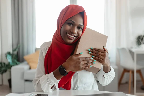快乐的非洲裔美国穆斯林妇女 头戴红色头巾 得到了她的分娩 拥抱盒和微笑在相机 家庭内部 Covid 19大流行病期间购物 — 图库照片