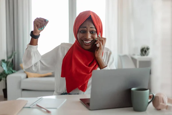 New job opportunity. Overjoyed african american muslim woman talking on cellphone and celebrating success, sitting with laptop at workplace at home office