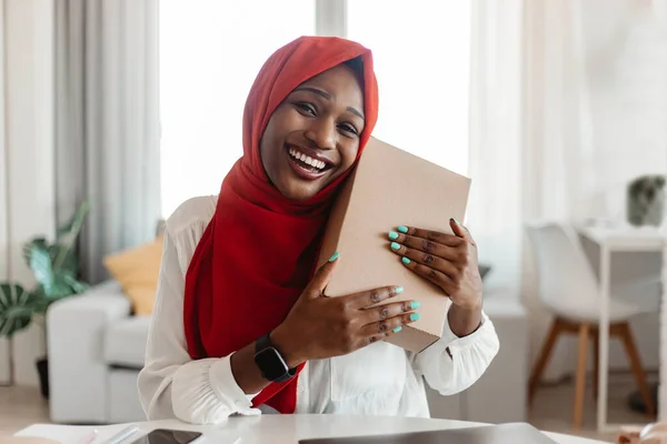 Contented buyer, shopaholism and commerce concept. Happy african american lady in hijab hugging cardboard box after receiving delivered parcel with products and smiling at camera