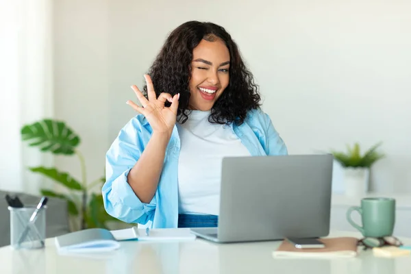 ラップトップジェスチャーを使用して陽気な黒人ビジネスマンOk現代のオフィスでカメラに座って笑顔をポーズウェブサイトを承認します 職場の屋内でオンラインで働くOkサインを示す女性 — ストック写真