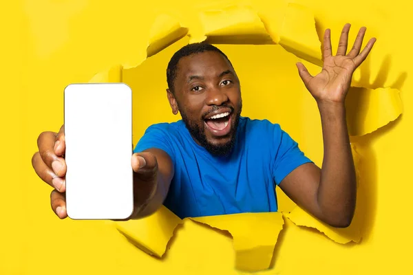 Overjoyed black man with open mouth showing smartphone with blank screen through torn hole in yellow paper, mockup. Great offer, ad, app and new device