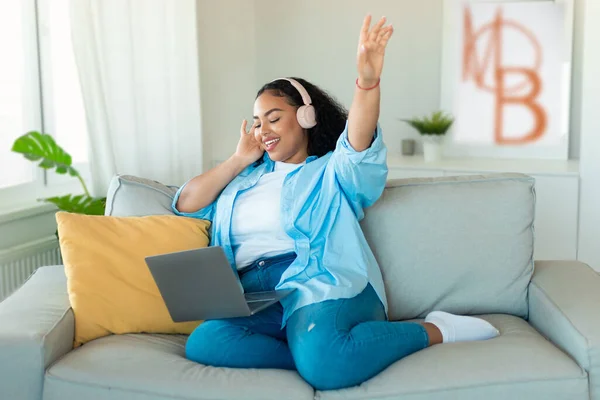 ラップトップコンピュータを使用してワイヤレスヘッドフォンを着用し 週末に自宅でソファに座って楽しむ音楽をオンラインで聴く幸せな黒人女性 お気に入り Song Concept — ストック写真