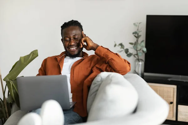 Home office concept. Happy black man working on laptop while sitting on comfy sofa at home interior, copy space. Excited guy using computer for study and work