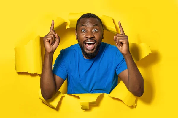 Portrait of surprised amazed black man pointing fingers up, standing in torn paper hole and looking at camera through breakthrough of yellow background.
