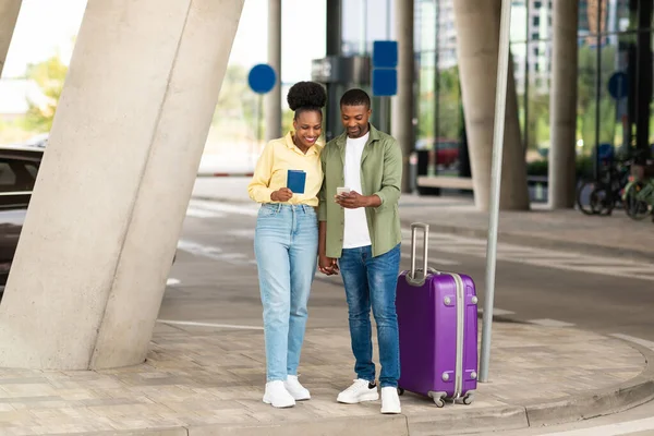 Travel Application. Happy African American Tourists Couple Using Smartphone Buying Flight Tickets Online Standing With Suitcase Near Airport Outdoors. Tourism And Mobile Technology Concept