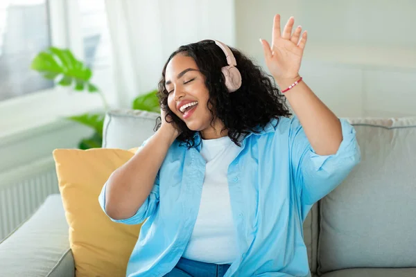 アフリカ系アメリカ人の陽気な女性は 家でソファに座って楽しむ音楽を聞いて 目を閉じて歌うイヤホンを身に着けています お気に入りの歌 素晴らしい音楽アプリケーションコンセプト — ストック写真