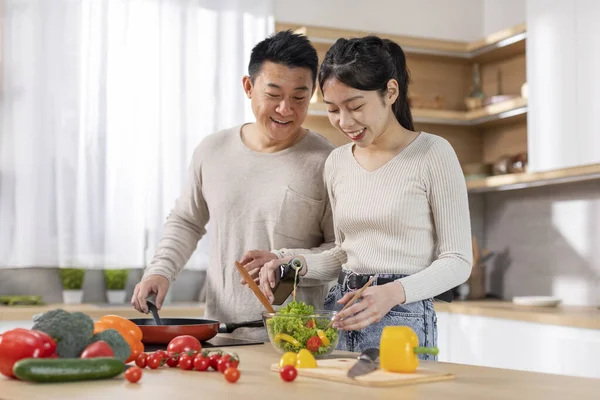 Beautiful asian couple enjoying cooking at home, happy chinese middle-aged man preparing healthy meal with his pretty young wife, making salad, cozy kitchen interior, copy space