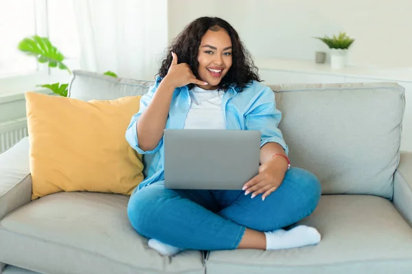 Happy African American Female Using Laptop And Gesturing Call Me Smiling To Camera Working And Communicating Online Sitting On Couch At Home. Freelance And Distance Communication