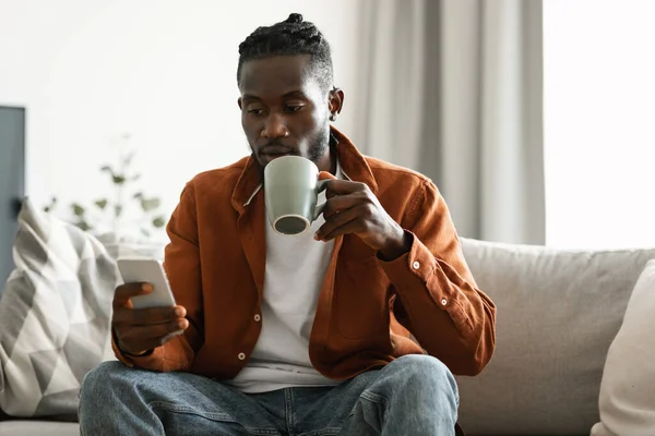 African american man using cool application on smartphone and drinking coffee, sitting on couch at home, copy space. Guy texting on cellphone. Mobile communication and technology