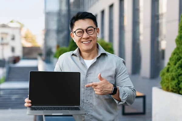 Portrait of korean businessman pointing at laptop computer with blank black screen, standing outdoors, mockup. People, technologies and remote work concept