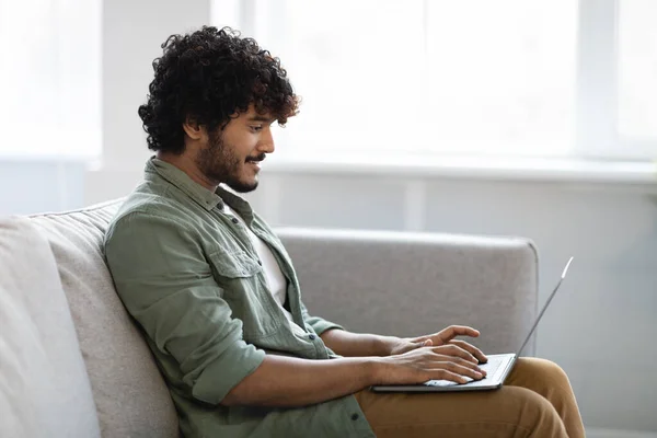 Side view of positive indian guy in casual outfit sitting on couch at home, using modern laptop, typing on computer keyboard and smiling, freelancer enjoying his remote job, copy space