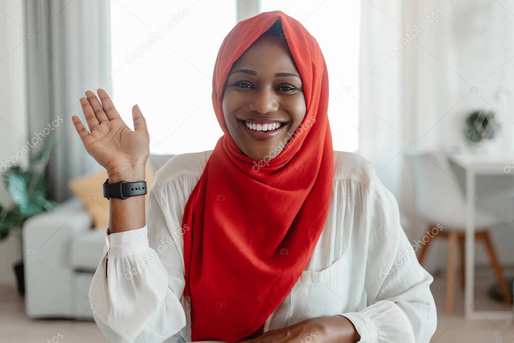 Greeting concept. Portrait of happy black muslim woman waving hello and smiling at camera, using laptop at home office. Positive female saying hi during video call