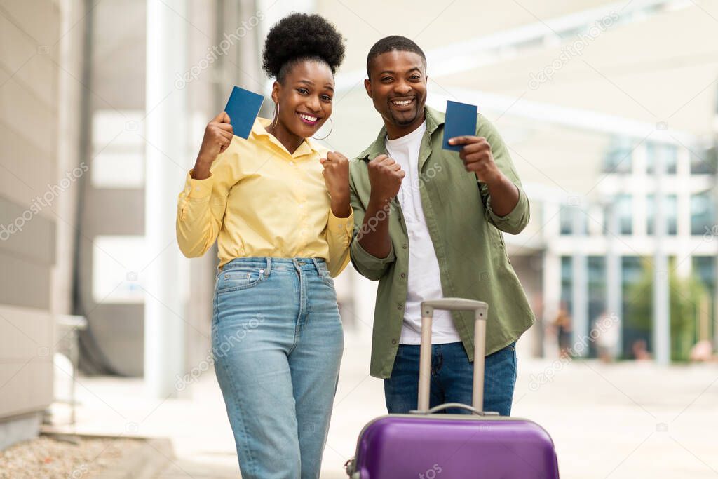Vacation, Finally. Joyful Black Travelers Couple Gesturing Yes Holding Passports Posing With Suitcase Smiling To Camera Standing Near Airport Outdoors. Great Travel And Tourism Offer Concept