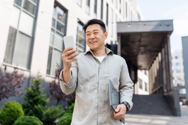 Orta yaşlı gülümseyen Asyalı işadamı, bilgisayarında akıllı telefonla yazı yazıyor, modern ofis binasının yanında yürüyor, açık havada. Yönetici ve başarılı iş, aygıtla uzaktan çalışma