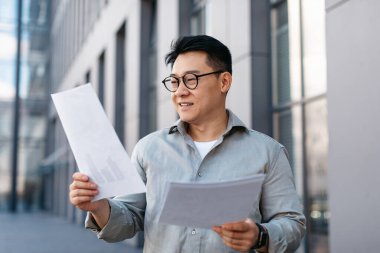 Belgelerle çalışan kendine güvenen Asyalı işadamı ofis binası yakınlarında açık havada raporlar okuyor. Liderlik ve proje analizi, CEO müdürü, patron ve sözleşme konsepti.
