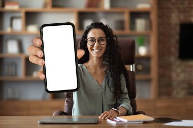 Pozitif çekici, kıvırcık, orta doğulu genç bir kadın çalışma masasında dizüstü bilgisayarı ve not defterleriyle oturuyor. Akıllı telefonu beyaz ekranlı, ofis içi, maketi, kopyalama alanı olan akıllı telefonu gösteriyor.