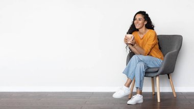 Neşeli, çekici, orta çağ ortadoğulu bir kadın, rahat bir kıyafetle evde koltukta oturmuş, bitki çayı ya da kahve içiyor, fotokopi odasına bakıyor ve gülümsüyor, beyaz arka plan, panorama.