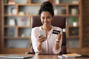 Pozitif genç iş kadını ofisten alışveriş yapıyor, çalışma masasında oturuyor, modern akıllı telefonu kullanıyor, internetten mal ve hizmetleri ödüyor, hafta sonu gezisi için bilet alıyor, fotokopi çekiyor.