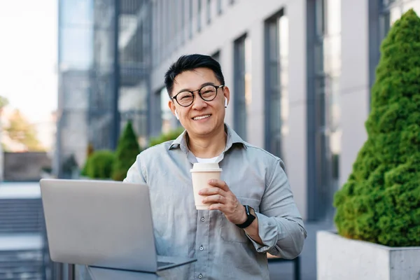 Happy asian middle aged businessman working on laptop and drinking coffee outdoors near office building, smiling at camera. Business outdoor, coffee break, lifestyle and digital nomad