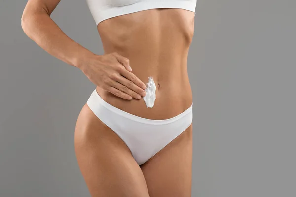 Unrecognizable young female in white underwear applying moisturising body lotion on belly, slim woman with sporty figure noirishing her skin with cream while standing over grey background, copy space