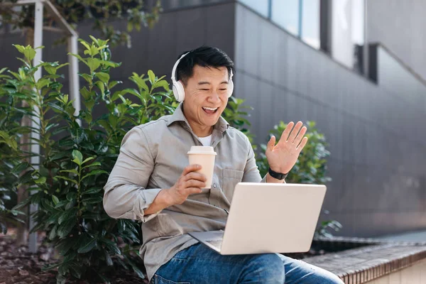 Excited asian middle aged businessman working on laptop outdoors, wearing headphones and having video call, drink coffee on bench near office building. Business outdoor, coffee break