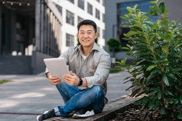 Szczęśliwy Azjatycki Przedsiębiorca Rozmawiający Tablecie Świeżym Powietrzu Siedzący Ławce Pobliżu — Zdjęcie stockowe