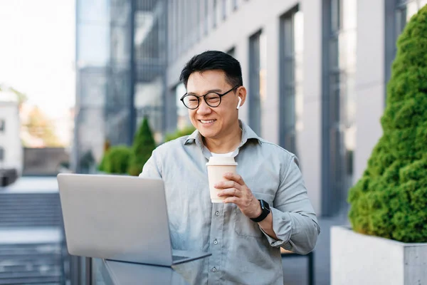 Happy mature asian man freelancer working on laptop and drinking coffee near office building outdoors. Business, coffee break, new normal, lifestyle and digital nomad