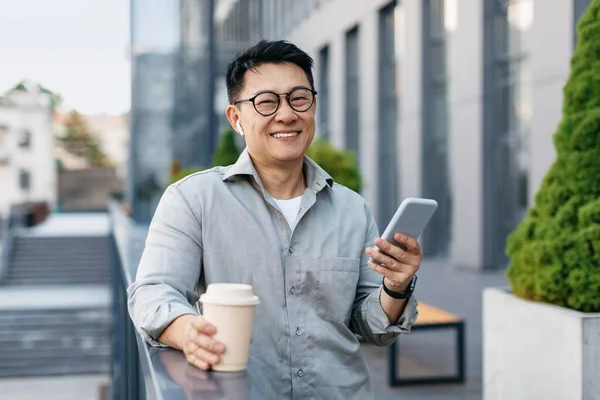 App for business and work. Happy asian male freelance worker with coffee takeaway, using smartphone and smiling at camera, standing near modern office building