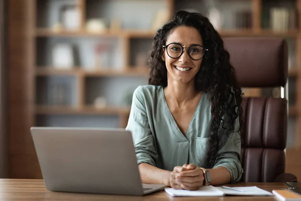 Cheerful brunette millennial woman in eyeglasses using laptop at office, lady sitting at workdesk in front of computer, smiling at camera, copy space. Career, business opportunities for millennials