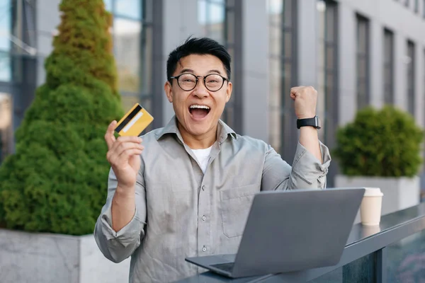 Overjoyed asian middle aged businessman holding credit card in front of laptop and making YES gesture while sitting near office building outdoors. Cool online promo concept