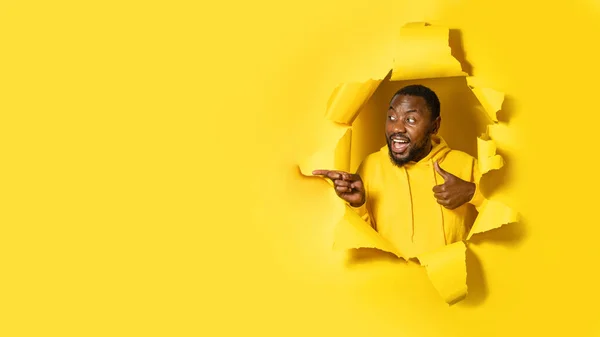 Amazed black man looking and pointing aside at free space, showing thumb up, posing through hole in yellow paper, panorama. Surprise expression, offer, sale, promo concept