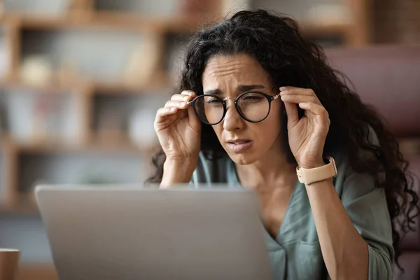 Millennial woman in casual outfit sitting at workdesk in front of laptop and touching her eyeglasses, suffering from sore eyes while working at office, copy space, closeup shot. Computer eyestrain