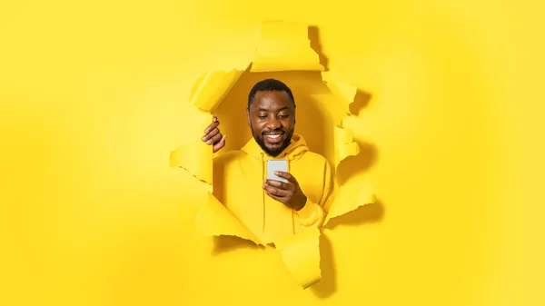 快乐的非洲裔美国人 拿着手机笑着 站在撕破的纸孔里 摆出一副黄色背景 全景的神气十足的样子 — 图库照片
