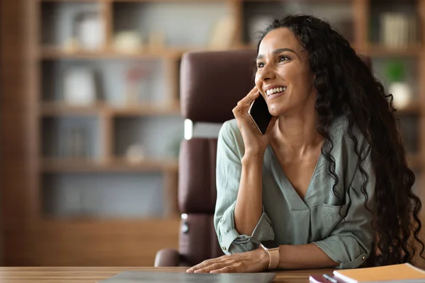 Happy pretty millennial woman in casual outfit entrepreneur sitting at table at office, having business conversation on phone and smiling, talking with business partner, looking at copy space