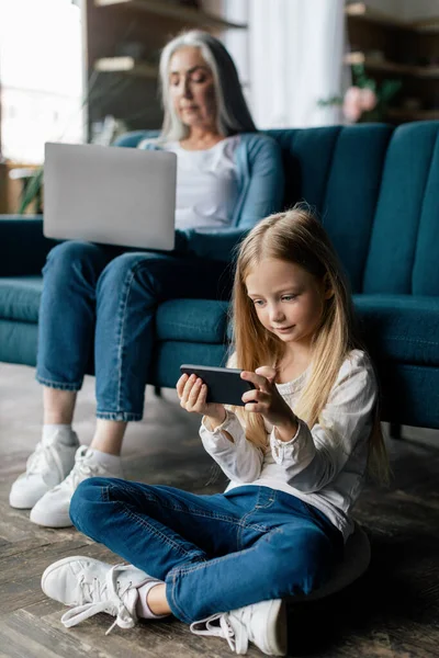 Busy European Little Granddaughter Plays Smartphone Elderly Grandmother Works Laptop — Stock Photo, Image