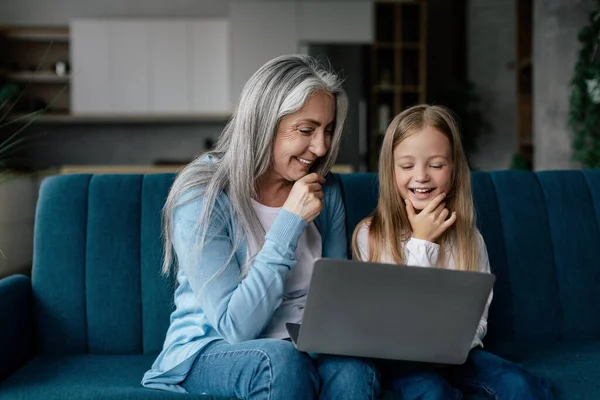 快乐的高加索小孙女和老奶奶想 在电脑上看视频 上网学习 在客厅里的沙发上玩游戏 由于Covid 在线通话 博客和在家里聊天 — 图库照片