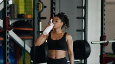 Susuzluk ve spor eğitimi. Genç, mutlu, Afro-Amerikan kadın şişeden su içiyor ve gülümsüyor spor salonunda sıkı çalıştıktan sonra dinleniyor, yavaş çekim, boş alan.